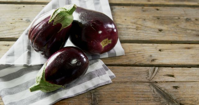 Fresh eggplants resting on a striped cloth with a rustic wooden background. Ideal for use in food blogs, recipe websites, kitchen-themed merchandise, and healthy eating campaigns.