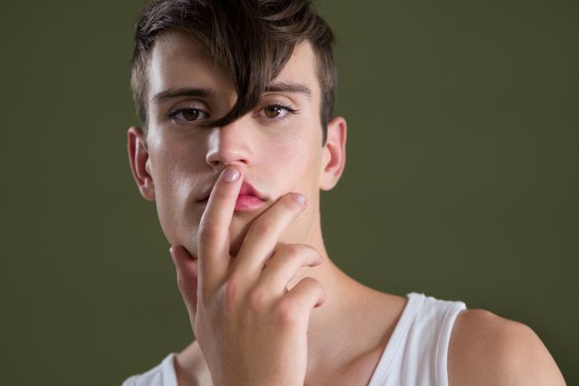 Androgynous man posing with finger on lips, wearing white tank top, against green background. Ideal for use in fashion editorials, modern lifestyle blogs, gender fluidity discussions, and promotional materials for contemporary clothing brands.