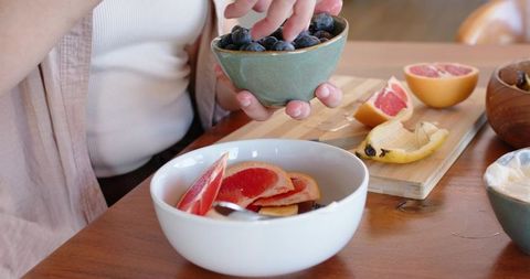 Woman adding blueberries to bowl with grapefruit slices on rustic breakfast table