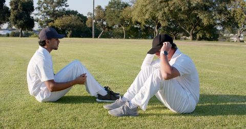 Diverse male teammates sitting on grass adjusting caps during sunny outdoor cricket practice