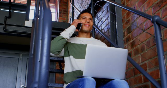 Man Sitting on Staircase Using Laptop and Talking on Phone in Industrial Setting - Download Free Stock Images Pikwizard.com