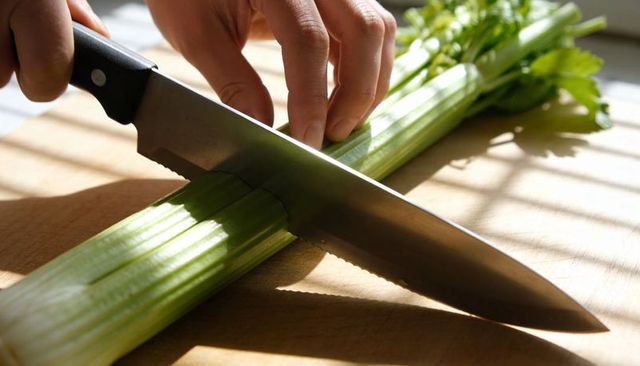 Closeup chef slicing celery on wooden cutting board with large knife in natural sunlight