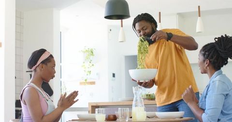 African family sharing sunlit kitchen meal while father tossing fresh green salad at table