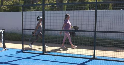 Women walking past padel court fence carrying paddles and racket bag in sunlight