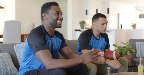 Friends watching football at home cheering together while holding football on couch