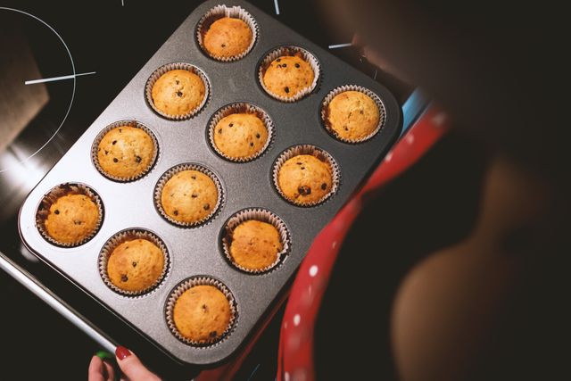 Freshly baked chocolate chip muffins in tray held by individual in kitchen. This can be used for content related to baking recipes, home baking, dessert ideas, and family cooking activities.