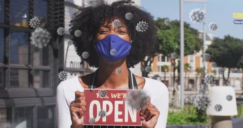 Small business owner holding we're open sign wearing blue mask with virus overlays