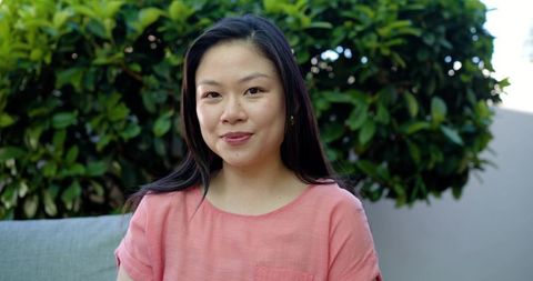 Smiling asian woman relaxing on patio sofa in coral blouse with lush green garden background