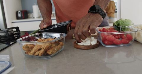 African american man prepping healthy lunches with grilled chicken, feta, tomatoes, kitchen counter