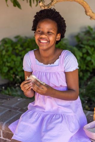 Sunlit smiling girl in pink pinafore holding sandwich while sitting on brick ledge in yard