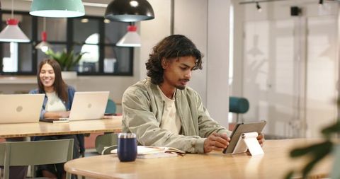Young man using tablet in modern coworking space with laptop and travel mug