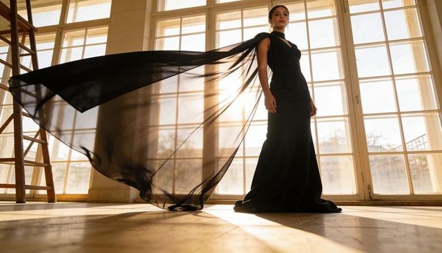 Model posing in black gown with flowing sheer cape backlit by sun in loft