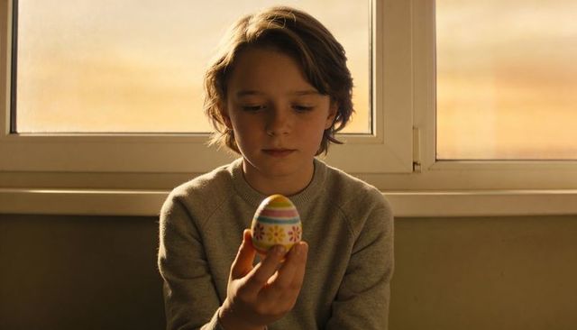 Contemplative boy holding decorated easter egg by window during golden hour warm light