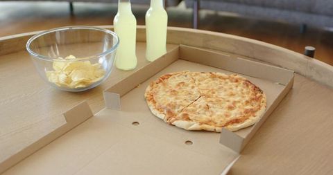Cheese pizza in open box on wooden coffee table with chips and lemonade, cozy living room