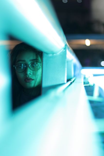 Young woman with glasses looks through railings bathed in blue neon light. Great for urban and modern themes, cyberpunk visual art, advertising for eyewear or tech products, or illustrating mystery and intrigue.