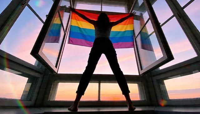 Silhouetted woman holding rainbow pride flag on window ledge at sunset, backlit