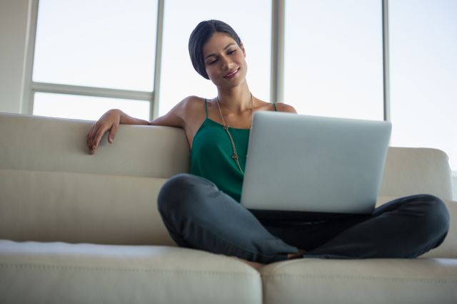 A businesswoman is sitting on a sofa in an office, using a laptop. She appears relaxed and is smiling, suggesting a comfortable and productive work environment. This image can be used for business, technology, and lifestyle content, illustrating modern work settings, remote work, or professional women in the workplace.