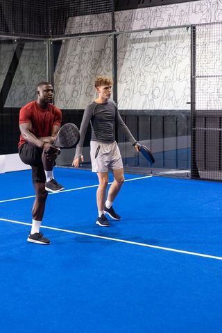 Young black and white teammates stretching on vibrant blue padel court holding rackets