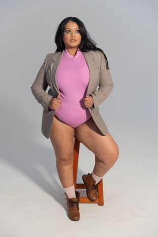 Confident mid-adult asian woman posing in taupe blazer and pink bodysuit on stool editorial