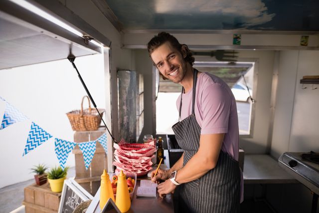 Young male waiter smiling while writing in a notepad at a food truck counter. He is wearing a casual outfit with an apron. Ideal for use in articles or advertisements about street food, small businesses, customer service, and outdoor dining experiences.
