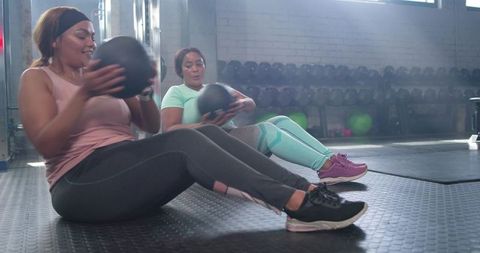 Diverse women doing medicine ball core workout together on gym floor, smiling and training
