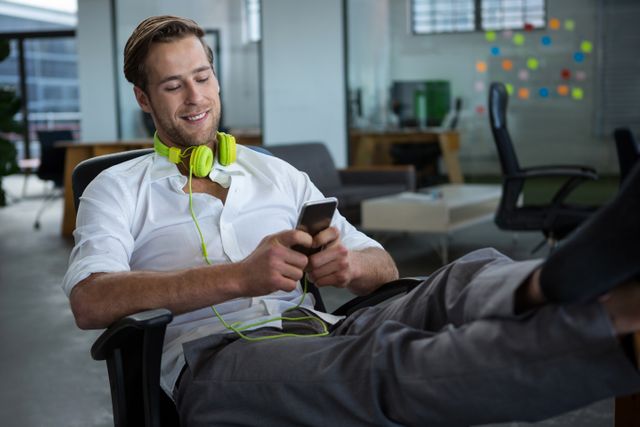 A young businessman is sitting in a modern office, smiling while using his mobile phone. He is wearing headphones and appears relaxed, with his feet up on the desk. This image can be used to depict a modern, casual work environment, technology use in business, or a relaxed corporate culture.