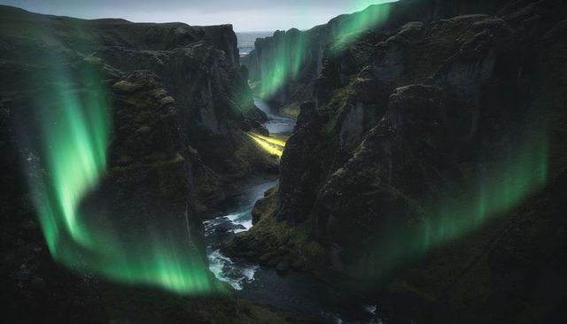 Aurora borealis flowing over mossy canyon, river carving toward distant sea, ledge glowing