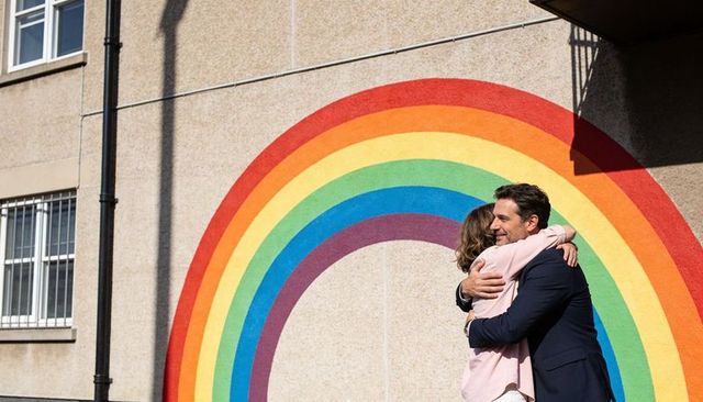 Couple hugging beside rainbow mural on urban wall, man in suit, woman in pink coat