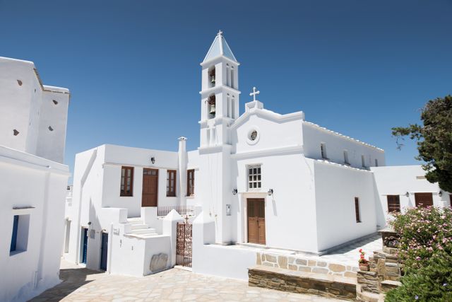 This image showcases a stunning white Orthodox church in a traditional Greek village. The crisp blue sky enhances the pristine white facades, embodying Mediterranean charm. Perfect for marketing travel destinations, promoting religious tourism, or illustrating articles about Greece and its architectural styles.
