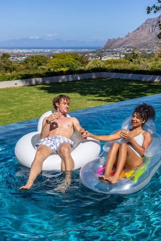 Mixed-race couple relaxing on pool floats holding drinks with ocean and mountain view