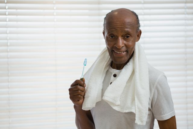 Senior man standing in bathroom holding toothbrush, smiling. Towel draped around neck, window with blinds in background. Ideal for promoting dental hygiene, senior health care, personal care products, and healthy lifestyle campaigns.