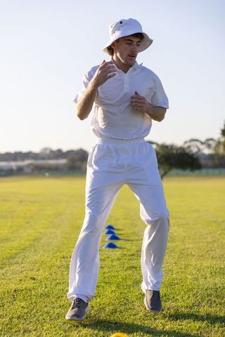 Male athlete training agile footwork in white uniform and bucket hat on sunny grass field