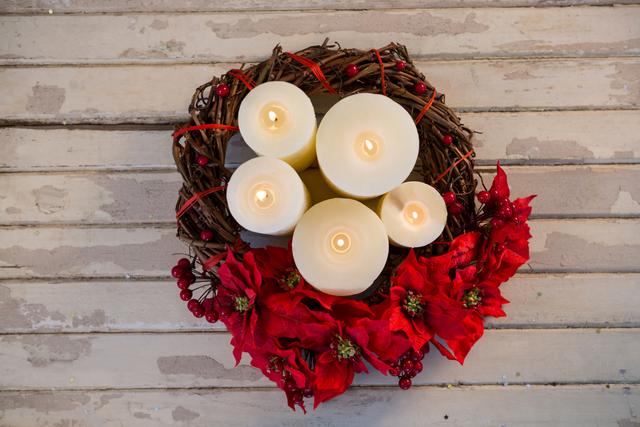 Top view of Christmas candles arranged in a wreath with red flowers and berries on a rustic wooden plank. Ideal for holiday greeting cards, festive invitations, seasonal blog posts, and home decor inspiration.
