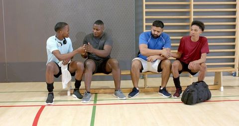 Diverse teammates chatting and clasping hands on gym bench after basketball practice