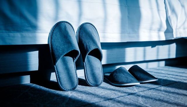 Soft dark slippers leaning and lying beside bed casting striped blinds shadows on carpet