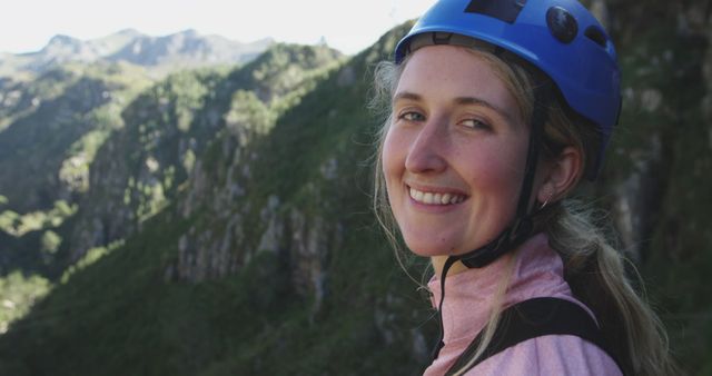 Woman is outdoors wearing climbing gear and helmet, smiling with mountains in background, suggesting adventure and enjoyment of nature. Perfect for promoting outdoor activities, adventure tourism, hiking, climbing expeditions, and nature travel plans.