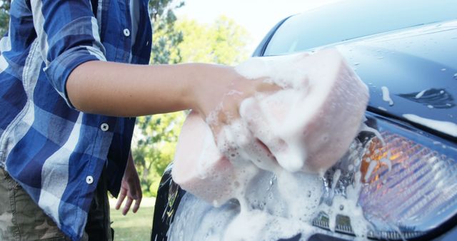 A young Caucasian male is washing a car, focusing on scrubbing the headlight with a soapy sponge, with copy space. Car washing is often a personal routine or a professional service to maintain vehicle cleanliness and appearance.