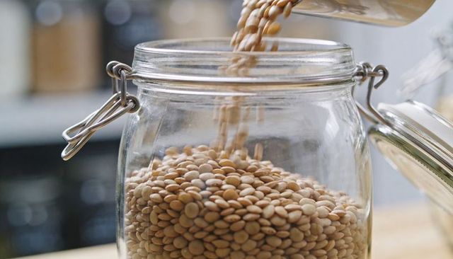 Pouring tan lentils into glass clip-top jar on kitchen countertop close-up for pantry