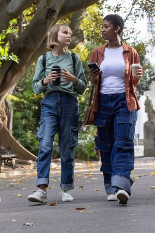 Two students walking on campus holding coffee and smartphone under autumn tree canopy