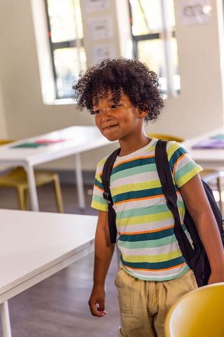 Elementary african american boy standing in classroom wearing striped shirt carrying backpack