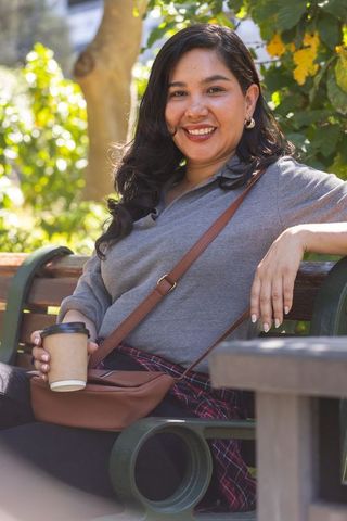 Smiling woman relaxing on park bench with takeaway coffee and crossbody bag, city lifestyle