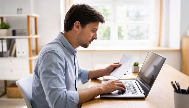 Remote worker typing on laptop while holding spiral notebook in bright home office