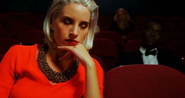 This photo shows a young woman with blond hair wearing a red dress and a large gold necklace, sitting alone in a nearly empty movie theater. The theater seats are a deep red color, and the background is fairly dark, with only a couple of other people distantly noticeable. This could be used to illustrate loneliness, boredom, movie-going experiences, or introverted moments. Ideal for articles about social experiences, entertainment industry, or personal reflection.