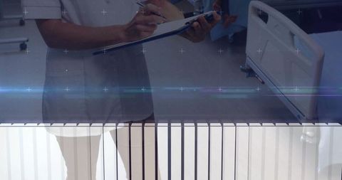 Clinician writing notes at hospital bedside holding clipboard and pen with visible bed rail
