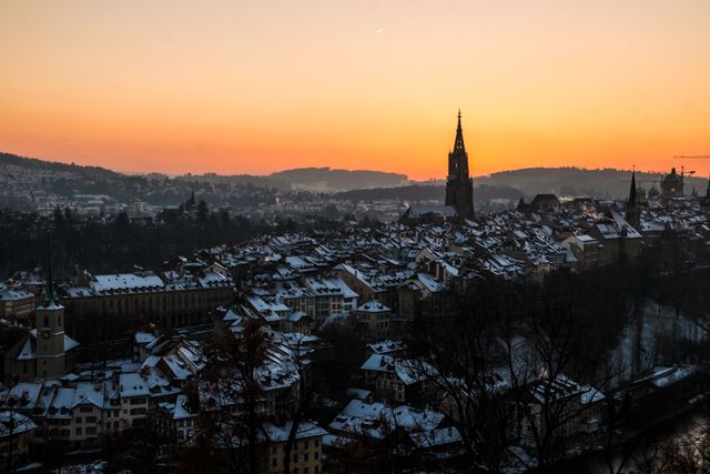 Snow-covered rooftops during sunset in a picturesque historic town with a prominent church spire, offering a serene and scenic winter scene. Ideal for travel magazines, winter-themed content, urban landscape presentations, and architectural interest pieces.