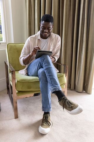 African american man using tablet while smiling in green armchair, sunlit living room