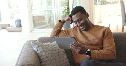 African American man reclining on couch using tablet in sunlit contemporary living room