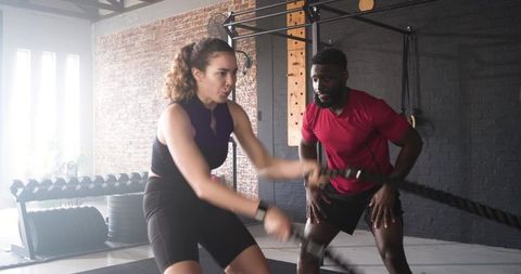 Diverse trainer and client exercising with battle ropes in gym