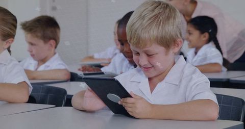 Blond boy engaging with tablet in classroom smiling while teacher observing