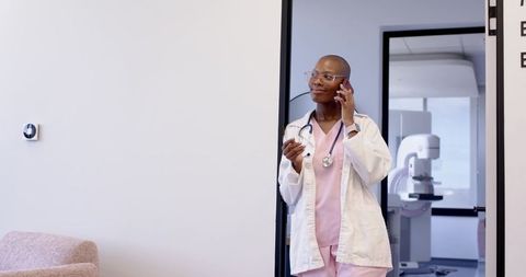 African female doctor talking on phone in clinic doorway in pink scrubs with stethoscope